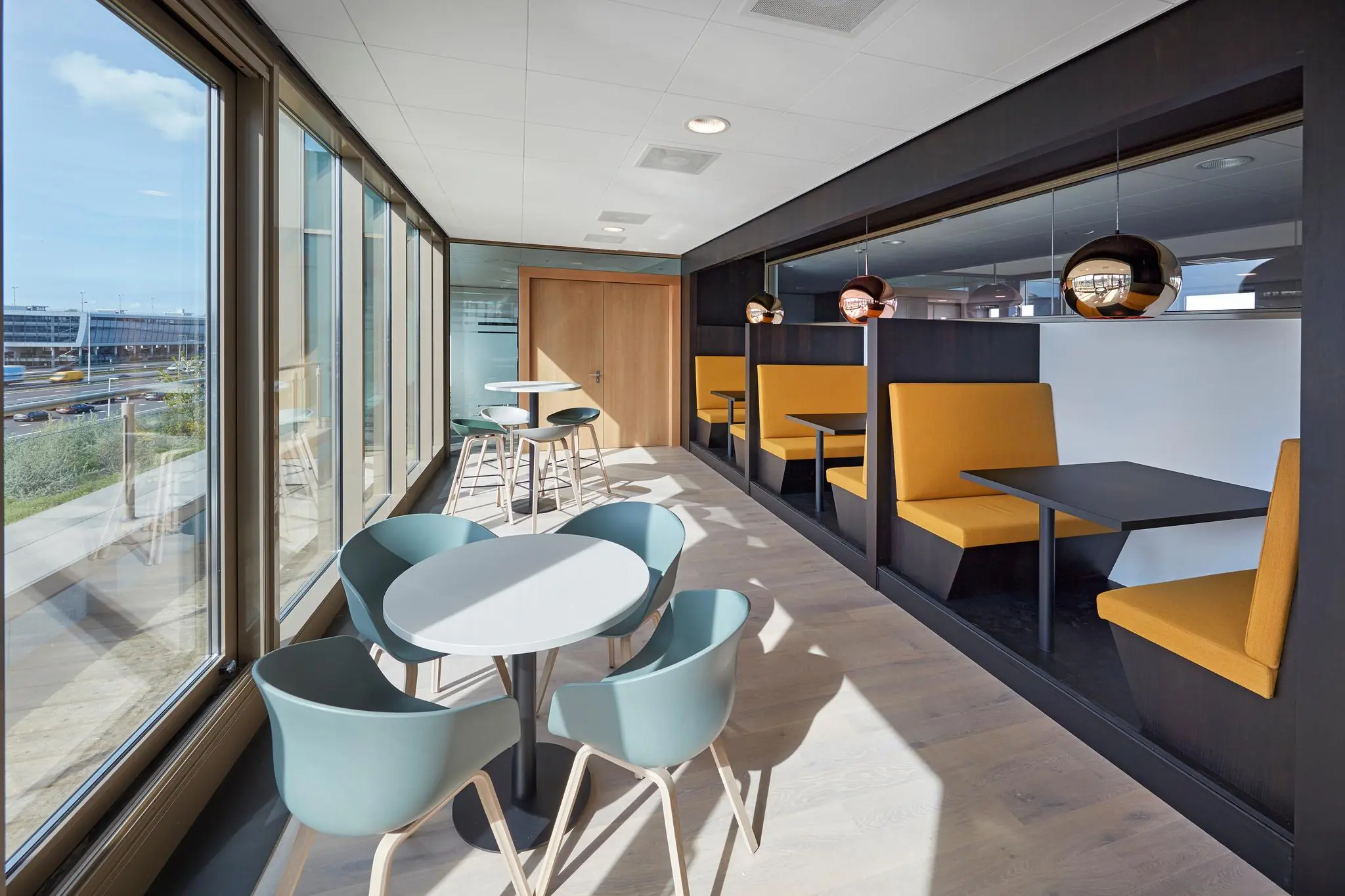 Moderne zitruimte in een kantoor aan de Fascinatio Boulevard met gele bankjes, ronde tafels en grote ramen met uitzicht op de weg.